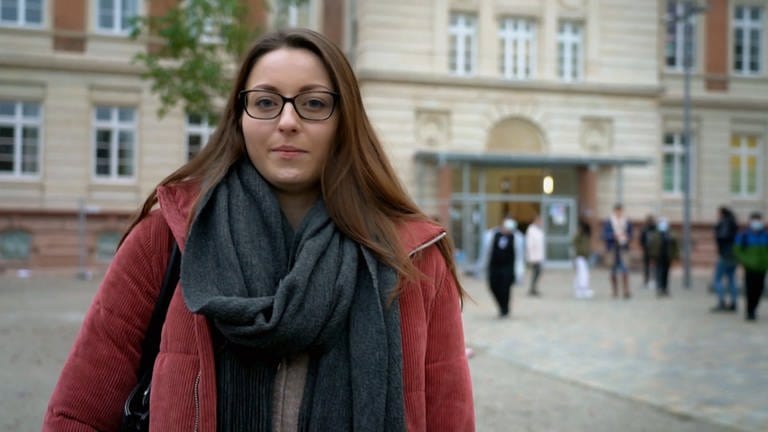 Eine junge Frau mit braunen, glatten Haaren und Brille schaut neutral in die Kamera