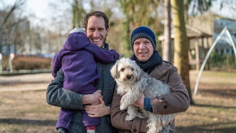 Zwei junge Männer halten ein Kleinkind und einen Hund auf dem Arm
