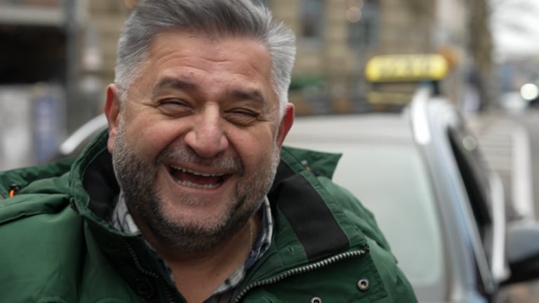 Iraklis ist Taxifahrer in Stuttgart. Er steht in der Stadt und lächelt herzlich in die Kamera. 