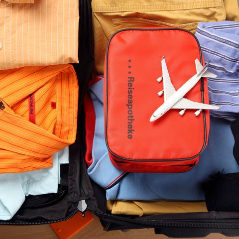 Eine Tasche mit der Aufschrift Reiseapothke liegt in einem gepackten Koffer. Auf ihr befindet sich ein kleines Flugzeug