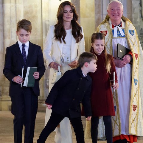 Eine Familie steht mit zwei Geistlichen vor einer Kirche, der kleine Junge in der Mitte pustet die Kerze seiner Schwester aus - Prinz Louis, Princess Charlotte