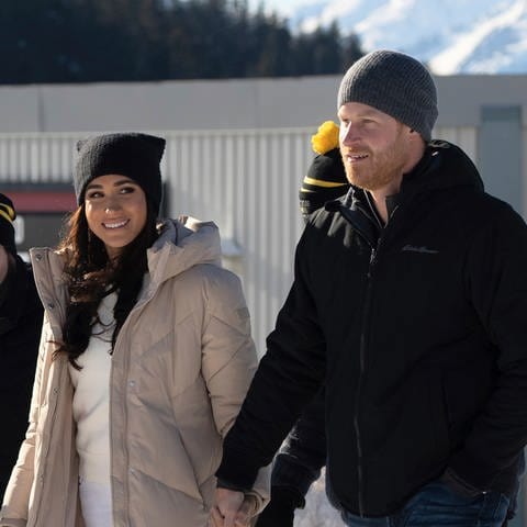 Prinz Harry und Herzogin Meghan auf einer Ski-Piste in Kanada. Sie trägt eine schwarze Mütze, unter ihrem beige-farbenen Mantel ist sie komplett in weiß gekleidet. Er trägt eine graue Mütze, eine schwarze Jacke und blaue Jeans. Die Sussexes besuchen das Traineingslager der Incivtus Games.