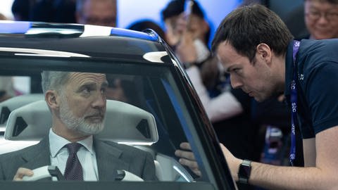 links sitzt ein grauhaariger Mann in einem sehr modernen silbernen Auto ohne Dach, König Felipe. Rechts neben dem Auto steht ein Mann heruntergebäugt und erklärt etwas.