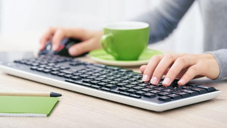 Eine Frau sitzt vor ihrem Computer und einer grünen Tasse und tippt in ihre Tastatur