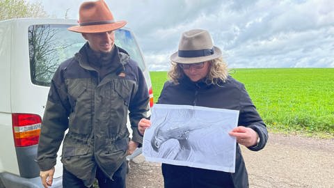 Zwei Menschen mit Hüten vor einer grünen Wiese in der Nähe von Bad Niedernau mit einer Schwarzweiß-Karte, auf der sich Gekände-Formationen erkennen lassen. 